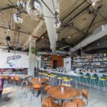 Spacious industrial-style restaurant with exposed ductwork, colorful mural, and a mix of booth and bar seating under spherical pendant lights.