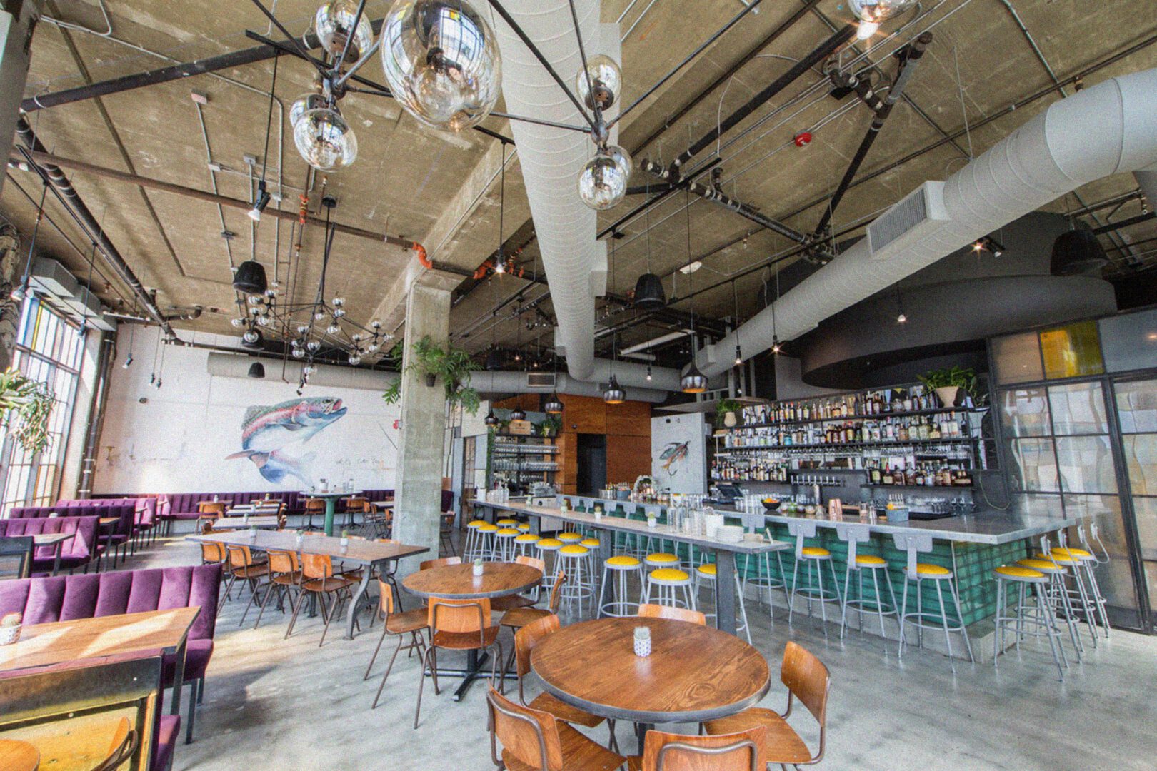 Spacious industrial-style restaurant with exposed ductwork, colorful mural, and a mix of booth and bar seating under spherical pendant lights.