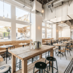 Modern, airy cafe interior with high ceilings, wooden tables, black metal chairs, and large windows letting in natural light.