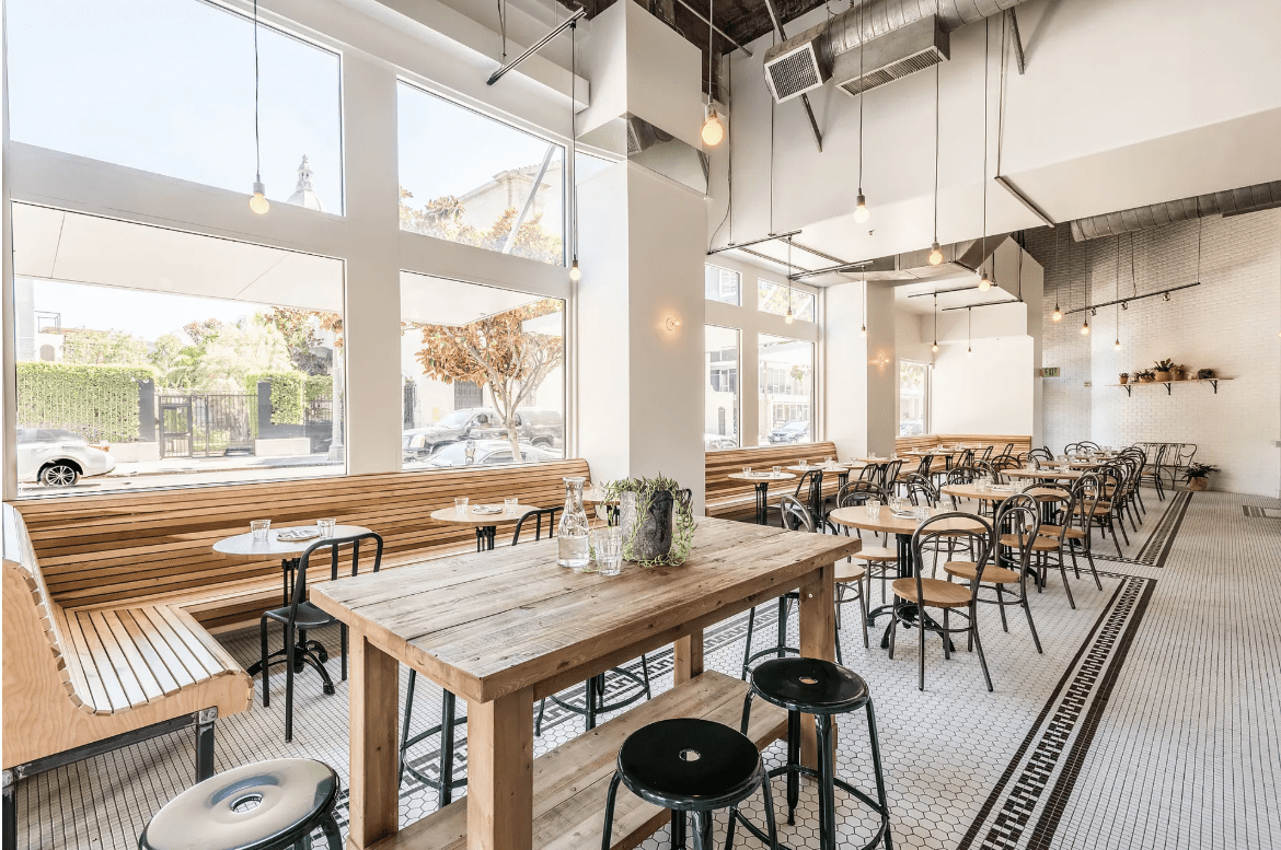 Modern, airy cafe interior with high ceilings, wooden tables, black metal chairs, and large windows letting in natural light.